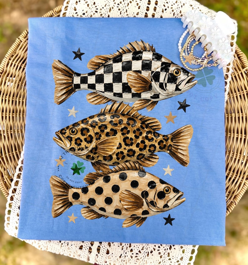 May include: A light blue decorative cloth featuring three stylized fish with unique patterns: a black and white checkerboard, a leopard print, and a spotted design. Black and gold stars are scattered around the fish. A white lace trim and a hair accessory are in the background.