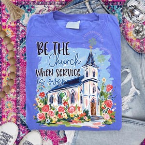 May include: A periwinkle blue t-shirt with the text "BE THE Church WHEN SERVICE is over" and a watercolor painting of a church surrounded by flowers. The design is centered on the shirt.