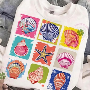 May include: White sweatshirt featuring a colorful seashell graphic. The design includes various seashell illustrations in pink, blue, orange, and yellow squares. The shirt has a crew neck and long sleeves, perfect for a casual, beach-themed look.