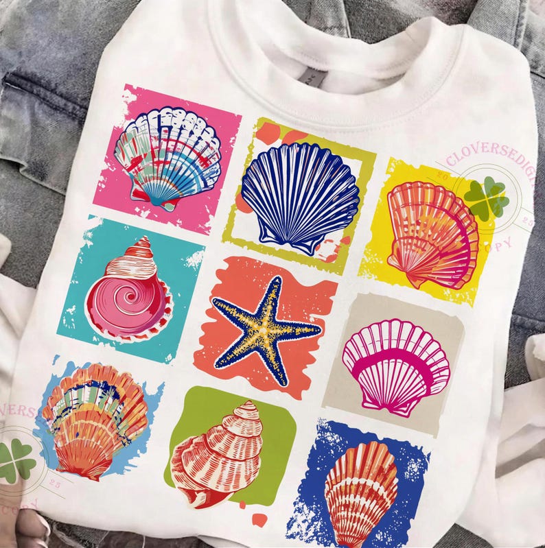 May include: White sweatshirt featuring a colorful seashell graphic. The design includes various seashell illustrations in pink, blue, orange, and yellow squares. The shirt has a crew neck and long sleeves, perfect for a casual, beach-themed look.