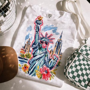May include: White t-shirt with a colorful illustration of the Statue of Liberty wearing sunglasses and blowing a bubble. The shirt also features the text "COMFORT COLORS" on the tag. The design includes flowers and New York City buildings.