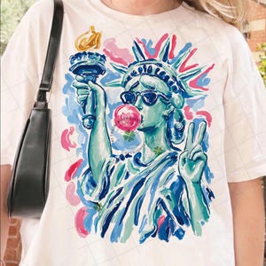 May include: Off-white t-shirt featuring a colorful illustration of the Statue of Liberty wearing sunglasses and blowing a bubblegum bubble. The Statue of Liberty is depicted in shades of blue and green, with pink and blue accents. The design includes the text "ALL RIGHTS RESERVED".