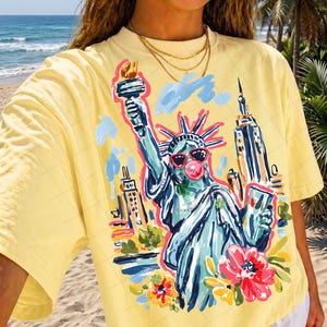 May include: Yellow t-shirt featuring a colorful illustration of the Statue of Liberty wearing sunglasses and blowing a bubblegum bubble. The design includes the New York City skyline and flowers. The shirt has short sleeves.