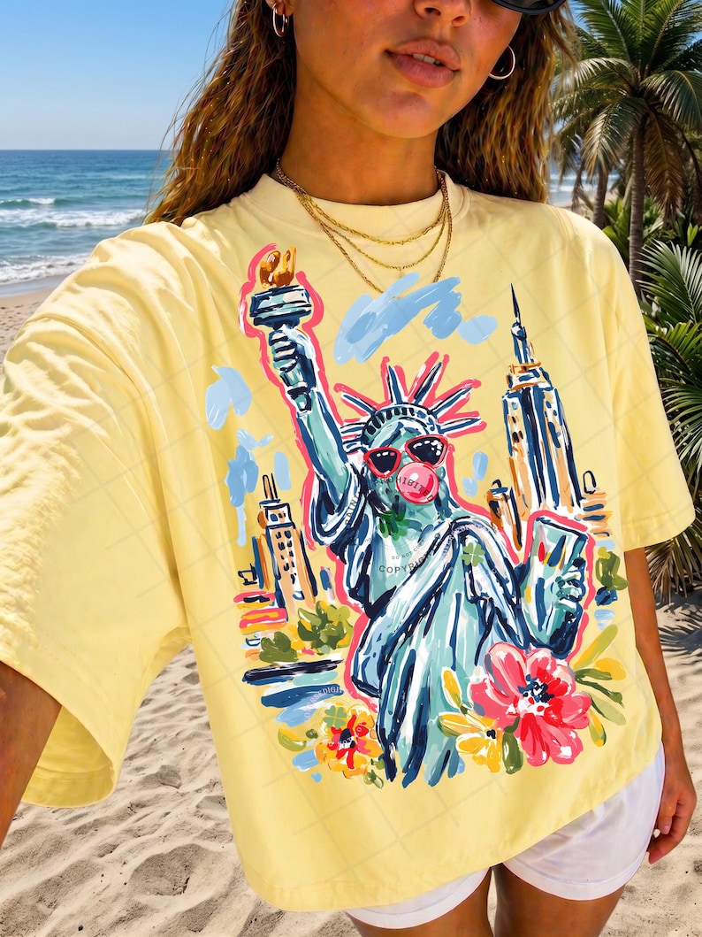 May include: Yellow t-shirt featuring a colorful illustration of the Statue of Liberty wearing sunglasses and blowing a bubblegum bubble. The design includes the New York City skyline and flowers. The shirt has short sleeves.