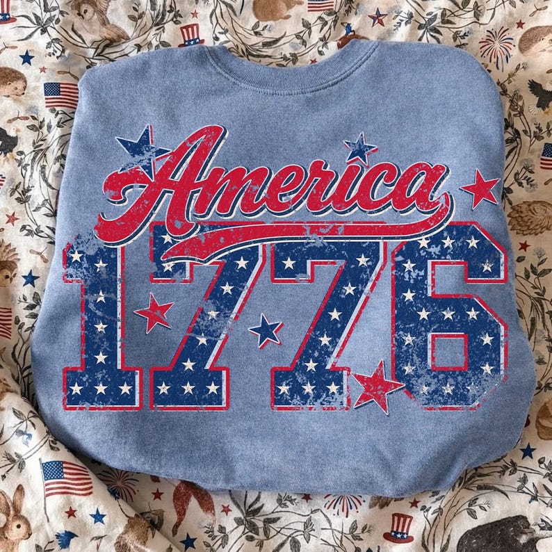 Może przedstawiać: Jasnoniebieska bluza z napisem "America" w czerwonym skrypcie i liczbą "1776" w kolorze niebieskim, ozdobiona białymi gwiazdami. Projekt ma postarzany, vintage wygląd, nawiązujący do patriotycznego motywu.