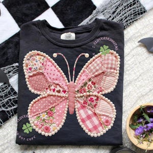 May include: Dark gray t-shirt featuring a large, patchwork butterfly design in shades of pink and floral patterns. The butterfly is outlined with pearls. The shirt has the text "Clover Colors" and "Clover Digital".