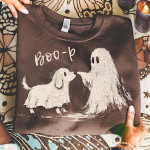May include: A dark brown crewneck sweatshirt featuring a distressed white graphic print of a small dog wearing a ghost costume standing next to a ghost. The text "BOO-p" is printed in a white, hand-lettered style above the two figures.