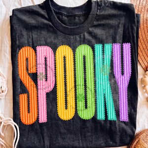以下が含まれることがあります： 黒の半袖Tシャツで、大きくカラフルな縦縞模様の文字で「SPOOKY」とプリントされています。各文字には特徴的なスカラップ状の縁取りがあり、オレンジ、ピンク、イエロー、ライムグリーン、ティール、パープルの順で並んでいます。