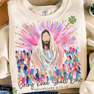 May include: Cream-colored sweatshirt with a watercolor illustration of Jesus with a colorful halo and crowd. The text "Every Knee Shall Bow" and "Philippians 2:10-13" are printed below the image. A clover logo is in the upper right corner.