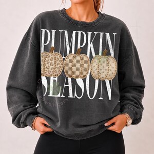 May include: A charcoal grey crewneck sweatshirt featuring the white text PUMPKIN SEASON. The design includes three illustrated pumpkins with distinct patterns: a leopard print, a checkered pattern, and a solid tan color with brown stems.