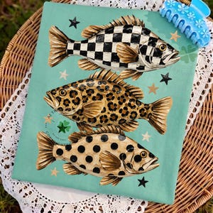 May include: A teal-colored decorative cloth featuring three stylized fish with unique patterns: a black and white checkerboard, leopard print, and black polka dots. Gold stars and a blue hair clip with floral details are also visible.