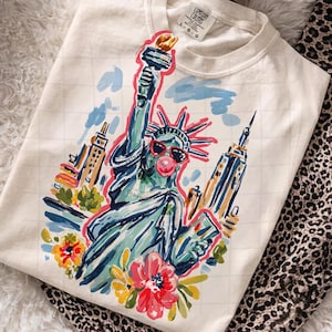 May include: Off-white sweatshirt featuring a colorful illustration of the Statue of Liberty wearing sunglasses and blowing a bubblegum bubble. The design includes New York City buildings and floral accents. The sweatshirt is made of a soft material.