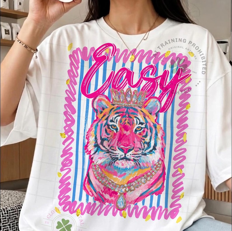 May include: White t-shirt featuring a colorful tiger illustration wearing a crown and necklace. The word "Easy" is written in pink script above the tiger. The design includes blue and white stripes and a pink border.