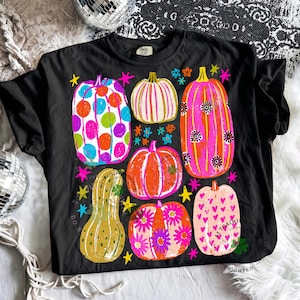 May include: A black t-shirt featuring a graphic print of six pumpkins in various shapes and colours, including polka dots, stripes, floral patterns, and hearts, accented with small colourful stars and flowers across the front of the garment.