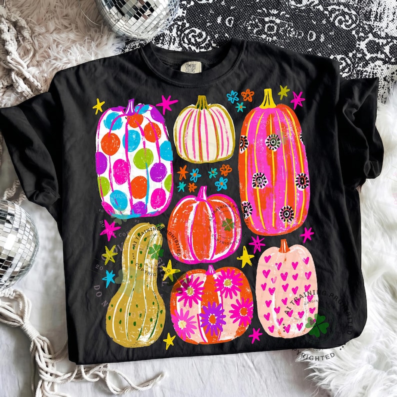 May include: A black t-shirt featuring a graphic print of six pumpkins in various shapes and colours, including polka dots, stripes, floral patterns, and hearts, accented with small colourful stars and flowers across the front of the garment.