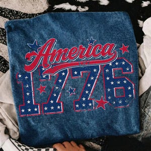 Może przedstawiać: Niebieska koszulka dżinsowa z napisem "America" w czerwonym skrypcie i liczbą "1776" w kolorze niebieskim, oba ozdobione białymi gwiazdami. Projekt nawiązuje do tematu patriotycznego.