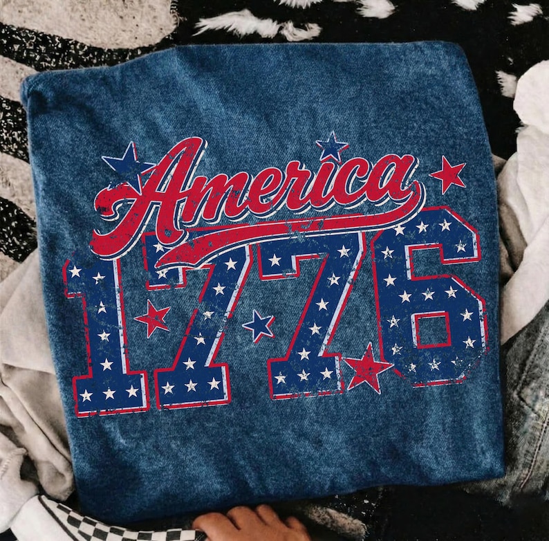 Może przedstawiać: Niebieska koszulka dżinsowa z napisem "America" w czerwonym skrypcie i liczbą "1776" w kolorze niebieskim, oba ozdobione białymi gwiazdami. Projekt nawiązuje do tematu patriotycznego.