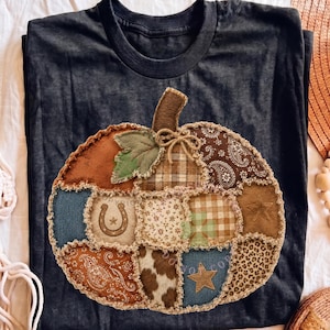 Könnte beinhalten: Ein dunkel anthrazitfarbenes T-Shirt mit einer großen, zentralen Kürbisgrafik aus verschiedenen Stoffflicken. Das Kürbisdesign umfasst braune Paisley-, Karomuster-, Leoparden-, Kuhfell- und Jeansstrukturen sowie ein Hufeisen und einen Stern.