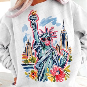 May include: White sweatshirt featuring a colorful illustration of the Statue of Liberty wearing sunglasses and blowing a bubblegum bubble. The design includes the New York City skyline and floral accents, creating a fun, artistic graphic.