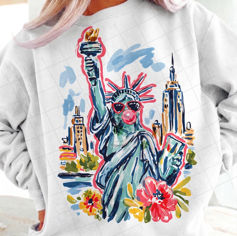 May include: White sweatshirt featuring a colorful illustration of the Statue of Liberty wearing sunglasses and blowing a bubblegum bubble. The design includes the New York City skyline and floral accents, creating a fun, artistic graphic.