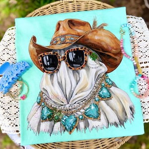 May include: A light turquoise t-shirt featuring a graphic illustration of a ghost wearing a brown cowboy hat, leopard print sunglasses, and a layered necklace with large teal heart-shaped gems and pearls. The design includes a small feather detail.