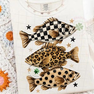 May include: White Comfort Colors t-shirt featuring three decorative fish with unique patterns: a black and white checkerboard, leopard print, and tan polka dots. The shirt is adorned with star and clover graphics.