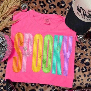 以下が含まれることがあります： 鮮やかなピンク色の半袖Tシャツで、大きなマルチカラーのストライプ文字で「SPOOKY」と書かれています。各文字はオレンジ、ピンク、イエロー、グリーン、ティール、パープルといった異なる色で構成されており、縁には装飾的なステッチが施されています。