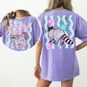 May include: A lilac-colored t-shirt featuring a raccoon graphic with the text "See Ya Soon". The design includes pink and teal stars and wavy lines. The raccoon has a black and white striped tail.