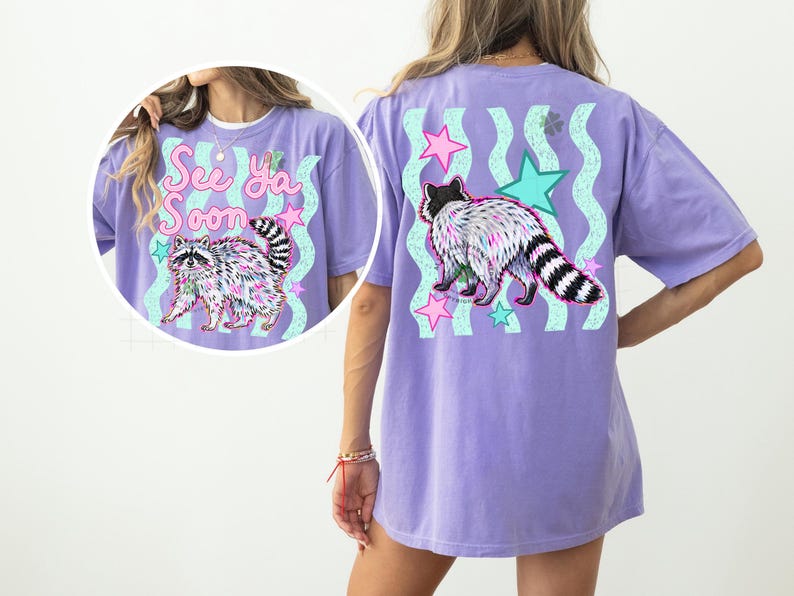 May include: A lilac-colored t-shirt featuring a raccoon graphic with the text "See Ya Soon". The design includes pink and teal stars and wavy lines. The raccoon has a black and white striped tail.