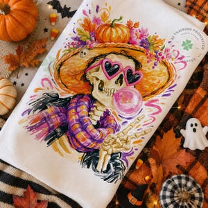 May include: A white kitchen towel featuring a colorful illustration of a skeleton wearing a large orange hat adorned with pumpkins and flowers, pink heart-shaped sunglasses, and a purple plaid scarf, while blowing a pink bubble gum bubble.
