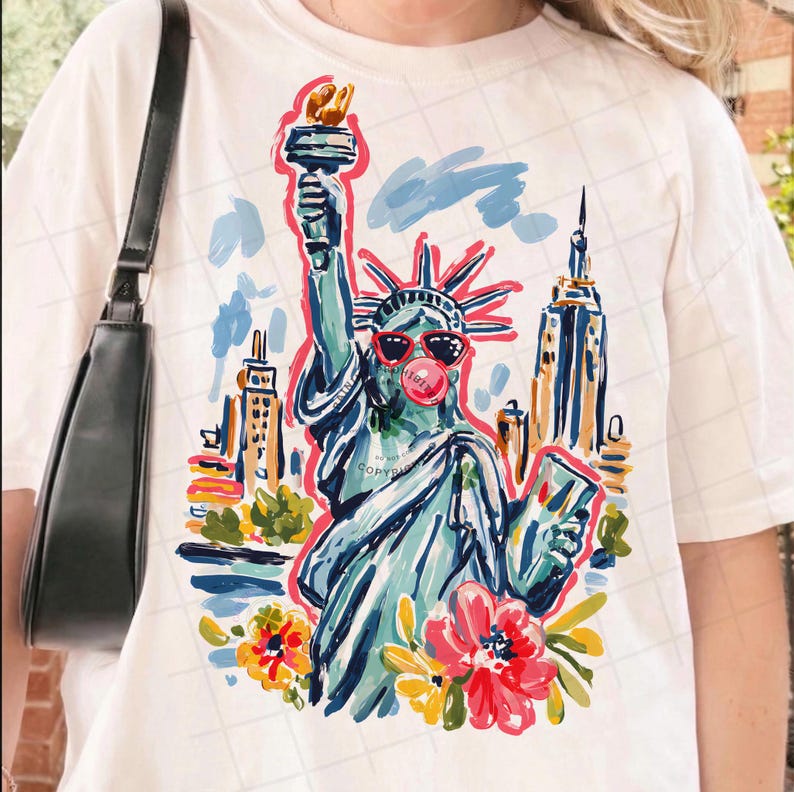 May include: White t-shirt featuring a colorful illustration of the Statue of Liberty wearing sunglasses and blowing a bubblegum bubble. The design includes New York City buildings and floral accents, creating a fun and vibrant graphic.