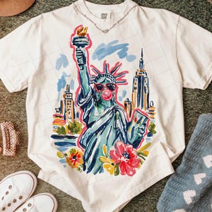 May include: Cream-colored t-shirt featuring a colorful illustration of the Statue of Liberty wearing sunglasses and blowing a bubble. The design includes the New York City skyline and flowers. The shirt is a Comfort Colors brand.