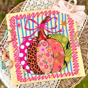 May include: A square fabric item featuring a central pumpkin illustration with varied patterns including leopard print, floral motifs, and polka dots. The pumpkin is topped with a bright pink bow against a background of blue vertical stripes and a pink border.