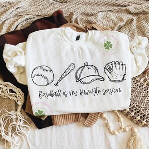 K&ouml;nnte beinhalten: Wei&szlig;es Sweatshirt mit einer schwarzen Strichzeichnung eines Baseballs, Schl&auml;gers, einer Kappe und eines Handschuhs. Der Text "Baseball is my favorite season" steht darunter. Das Sweatshirt liegt gefaltet auf einer neutralfarbenen Oberfl&auml;che.
