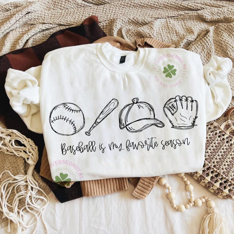 K&ouml;nnte beinhalten: Wei&szlig;es Sweatshirt mit einer schwarzen Strichzeichnung eines Baseballs, Schl&auml;gers, einer Kappe und eines Handschuhs. Der Text "Baseball is my favorite season" steht darunter. Das Sweatshirt liegt gefaltet auf einer neutralfarbenen Oberfl&auml;che.