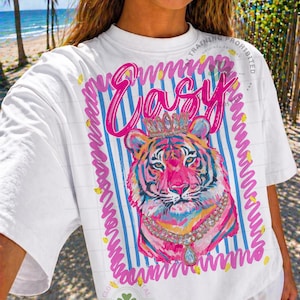 May include: White t-shirt with a colorful tiger graphic wearing a crown and necklace. The word "Easy" is written in pink script above the tiger. The design is framed with blue stripes and pink wavy lines.
