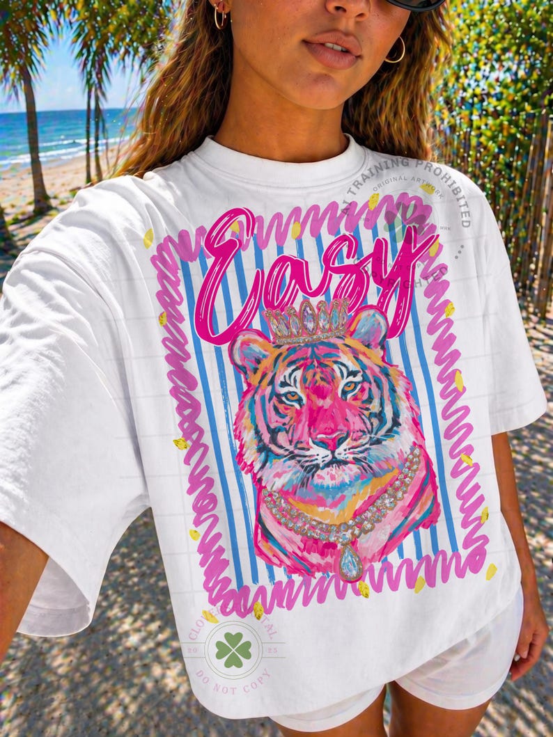 May include: White t-shirt with a colorful tiger graphic wearing a crown and necklace. The word "Easy" is written in pink script above the tiger. The design is framed with blue stripes and pink wavy lines.