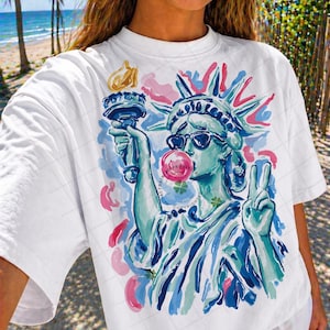 May include: White t-shirt featuring a colorful illustration of the Statue of Liberty wearing sunglasses and blowing a bubblegum bubble. The design includes pink, blue, and green accents. The Statue of Liberty is holding a torch and making a peace sign.