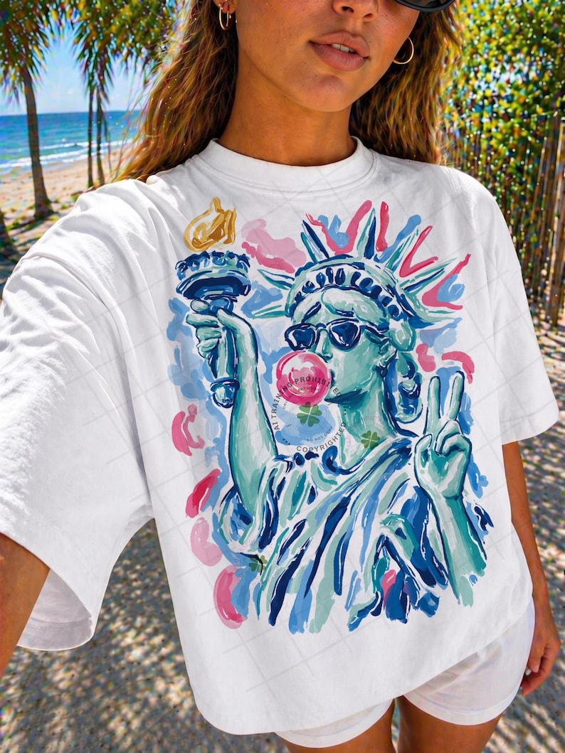 May include: White t-shirt featuring a colorful illustration of the Statue of Liberty wearing sunglasses and blowing a bubblegum bubble. The design includes pink, blue, and green accents. The Statue of Liberty is holding a torch and making a peace sign.