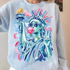 May include: Light blue sweatshirt featuring a colorful graphic of the Statue of Liberty wearing sunglasses and blowing a bubblegum bubble. The Statue of Liberty is holding a torch and making a peace sign. The sweatshirt has a crew neck and long sleeves.