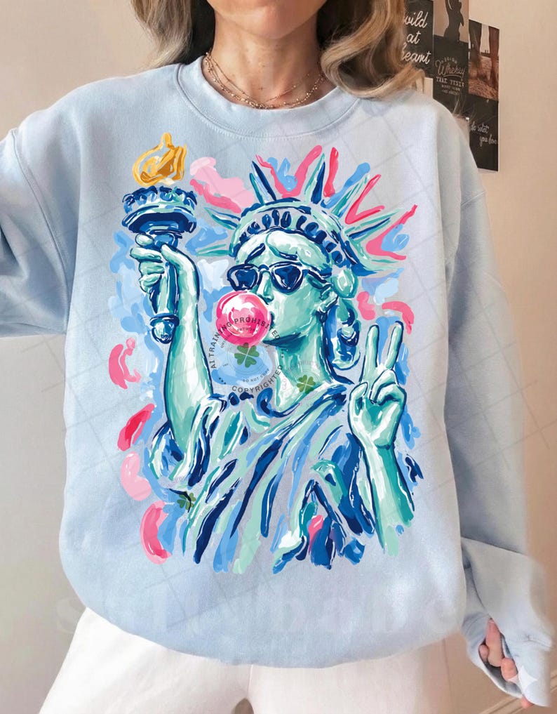 May include: Light blue sweatshirt featuring a colorful graphic of the Statue of Liberty wearing sunglasses and blowing a bubblegum bubble. The Statue of Liberty is holding a torch and making a peace sign. The sweatshirt has a crew neck and long sleeves.