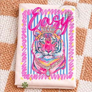 May include: A light pink kitchen towel with a colorful tiger illustration wearing a crown and a jeweled necklace. The word "Easy" is written in pink script above the tiger. Blue and pink stripes and squiggly lines frame the design.