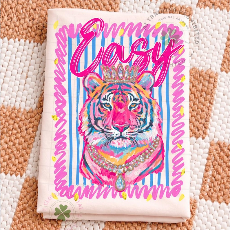 May include: A light pink kitchen towel with a colorful tiger illustration wearing a crown and a jeweled necklace. The word "Easy" is written in pink script above the tiger. Blue and pink stripes and squiggly lines frame the design.