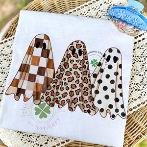 May include: A white t-shirt featuring a graphic of three ghosts. The first ghost has a brown and white checkered pattern, the second has a tan and brown leopard print, and the third has black polka dots. A light green clover logo is visible.