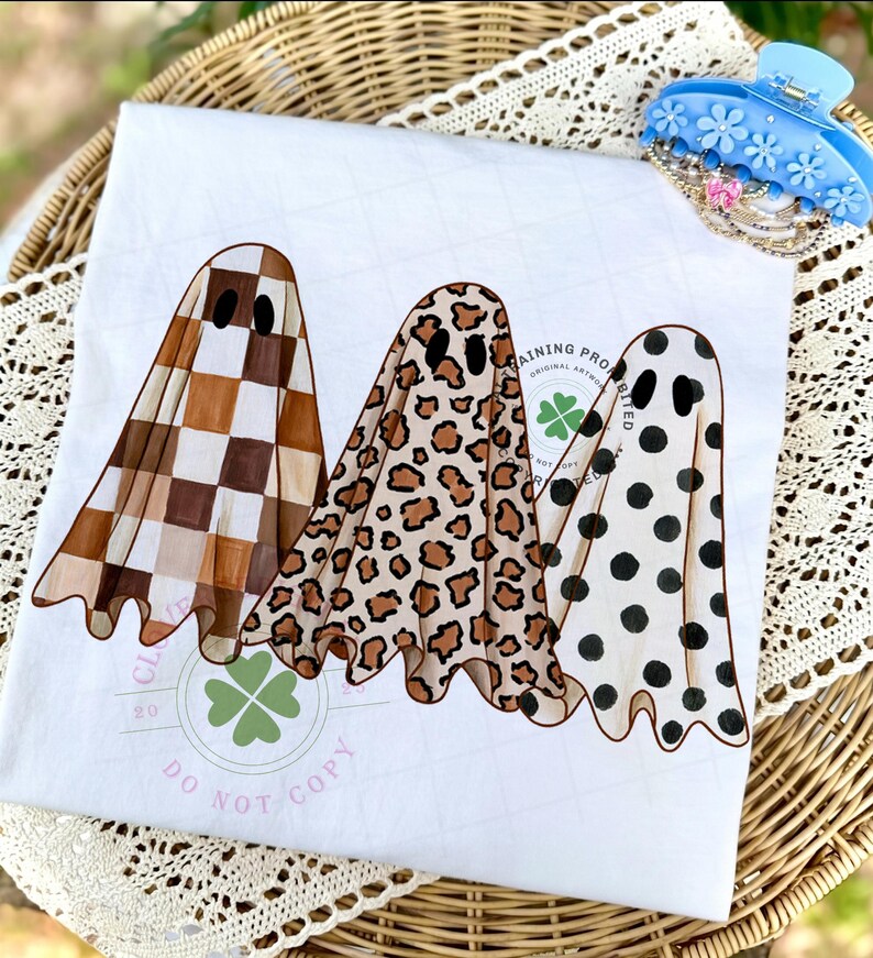 May include: A white t-shirt featuring a graphic of three ghosts. The first ghost has a brown and white checkered pattern, the second has a tan and brown leopard print, and the third has black polka dots. A light green clover logo is visible.