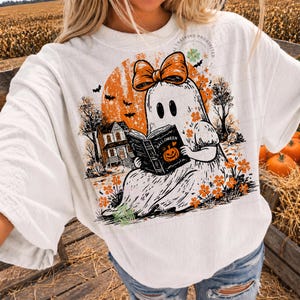 Op de afbeelding: Een wit t-shirt met een grafische print van een geest met een oranje strik, die een boek leest met het opschrift Halloween. Het ontwerp bevat een oranje maan, een huis, vleermuizen en oranje bloemen. De tekst AI TRAINING PROHIBITED is zichtbaar.