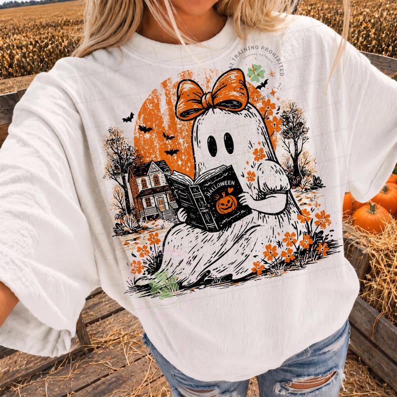 Op de afbeelding: Een wit t-shirt met een grafische print van een geest met een oranje strik, die een boek leest met het opschrift Halloween. Het ontwerp bevat een oranje maan, een huis, vleermuizen en oranje bloemen. De tekst AI TRAINING PROHIBITED is zichtbaar.