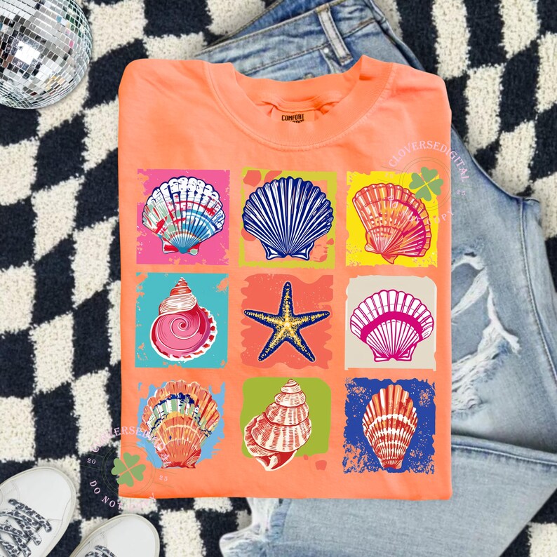 May include: Coral-colored t-shirt featuring a grid of colorful seashell illustrations. Each seashell is set against a different vibrant background, including pink, blue, and yellow. The shirt is laid flat on a denim surface.
