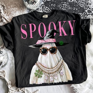 May include: A black t-shirt featuring the word SPOOKY in large pink letters. The graphic shows a white ghost wearing a black witch hat with a pink leopard print band, leopard print sunglasses, and multiple layered pearl and crystal necklaces.