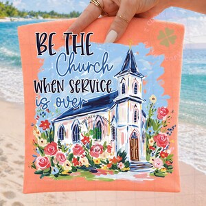 May include: Peach-colored t-shirt with the text "BE THE Church WHEN SERVICE is OVER" above a watercolor painting of a church surrounded by flowers. The church has a blue roof and a gold cross.